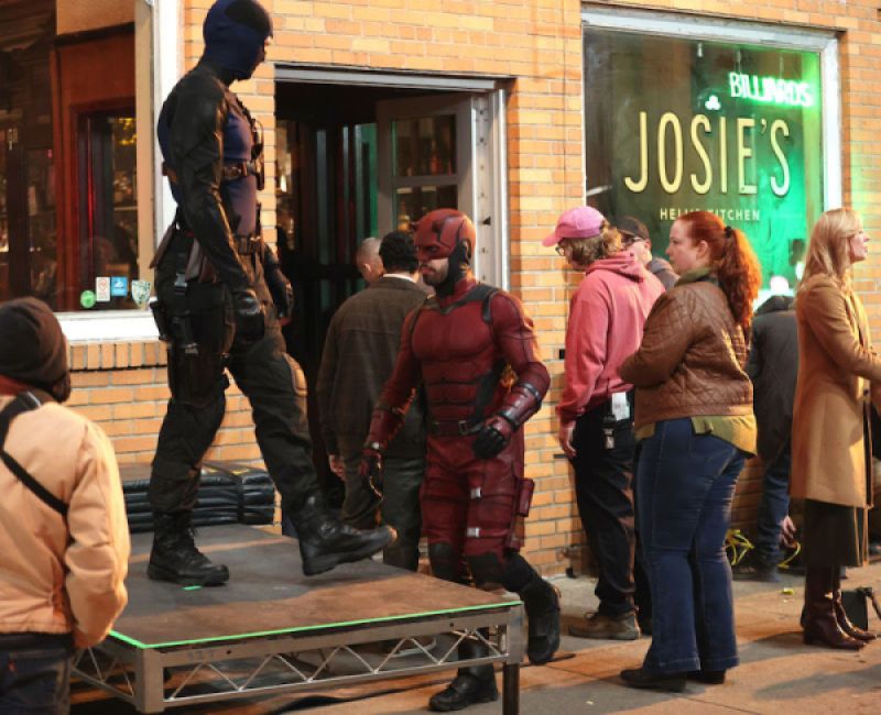 Matt Murdock as Daredevil, in a crowd of people outside a bar that has "Josie's" written in the window. Another figure in a black super suit is standing on a platform.