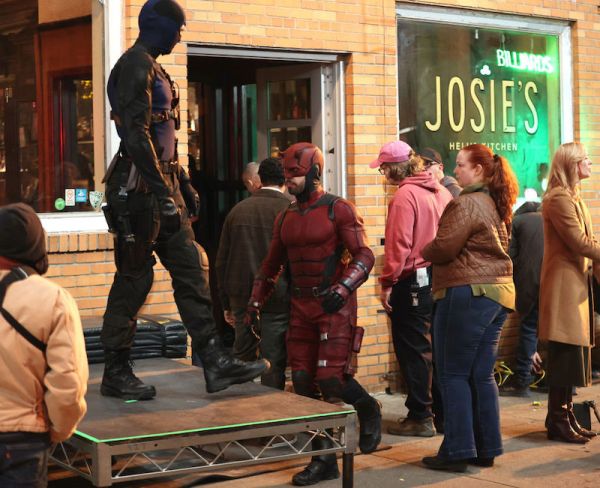 Matt Murdock as Daredevil, in a crowd of people outside a bar that has "Josie's" written in the window. Another figure in a black super suit is standing on a platform.