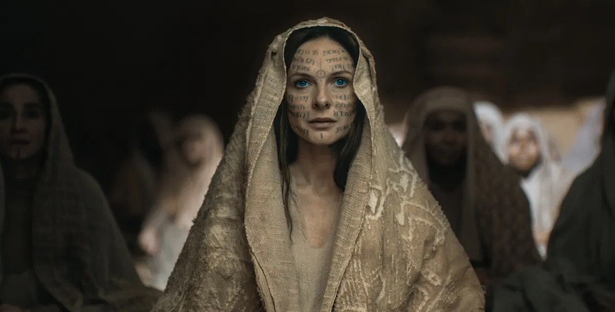 Rebecca Ferguson as Jessica in Dune: Part Two, wearing a gold shawl draped over her head. Her face is covered in writing.