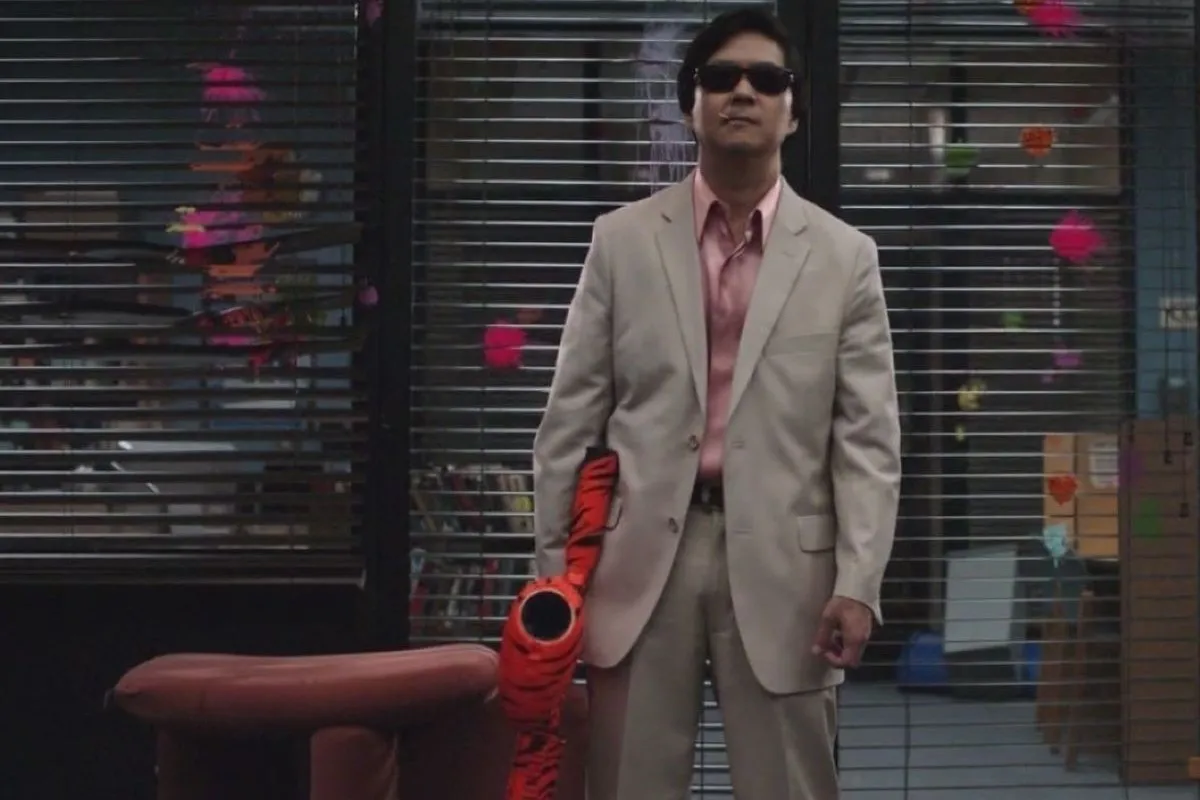 Still from Community episode Modern Warfare. Ken Jeong, a Chinese man in a grey suit and sunglasses, poses in front of a door with an orange and black striped paintball machine gun.