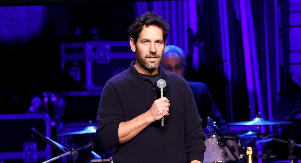 Paul Rudd holds a microphone on stage. Musicians are visible behind him.