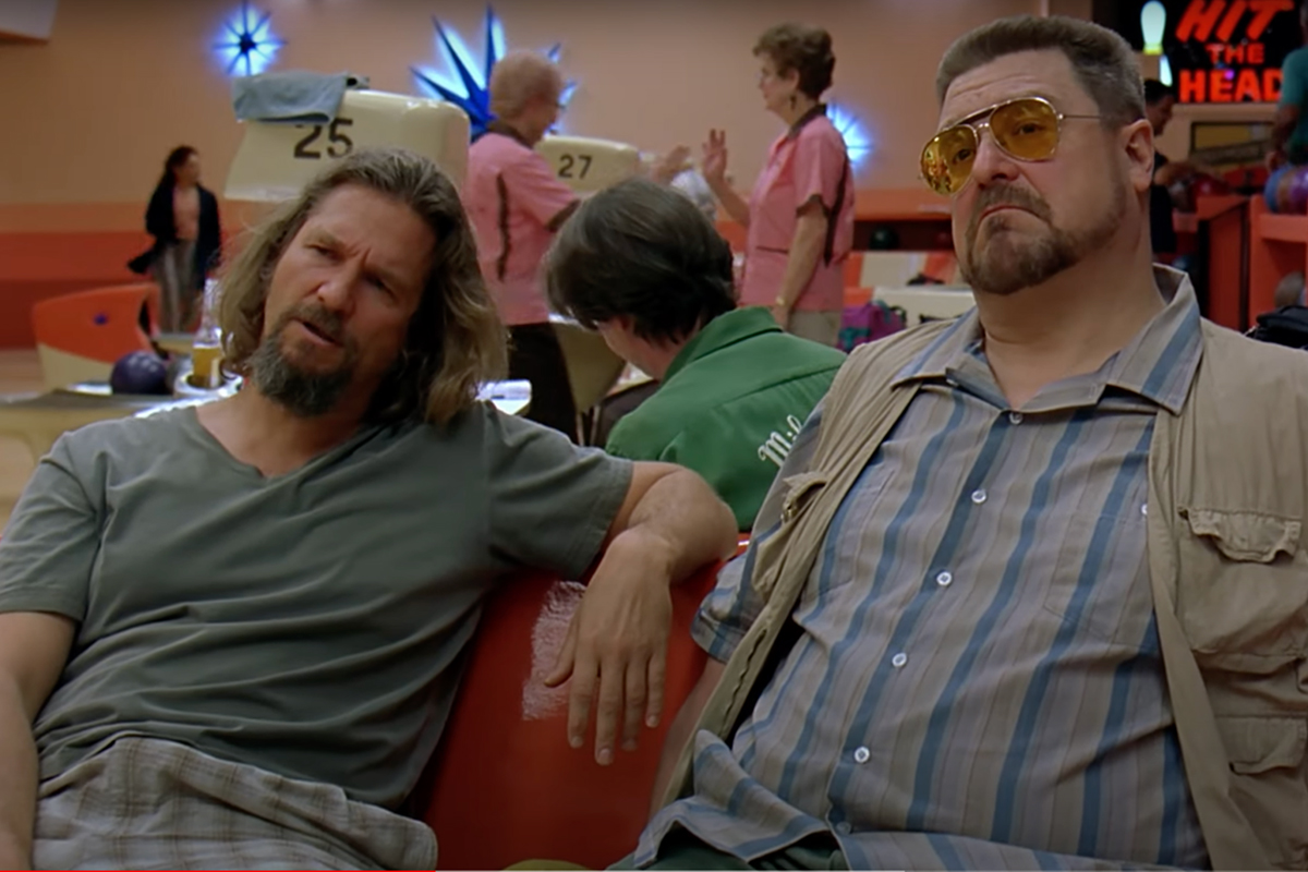Jeff Bridges and John Goodman leaning on the back of a shair in a bowling alley 