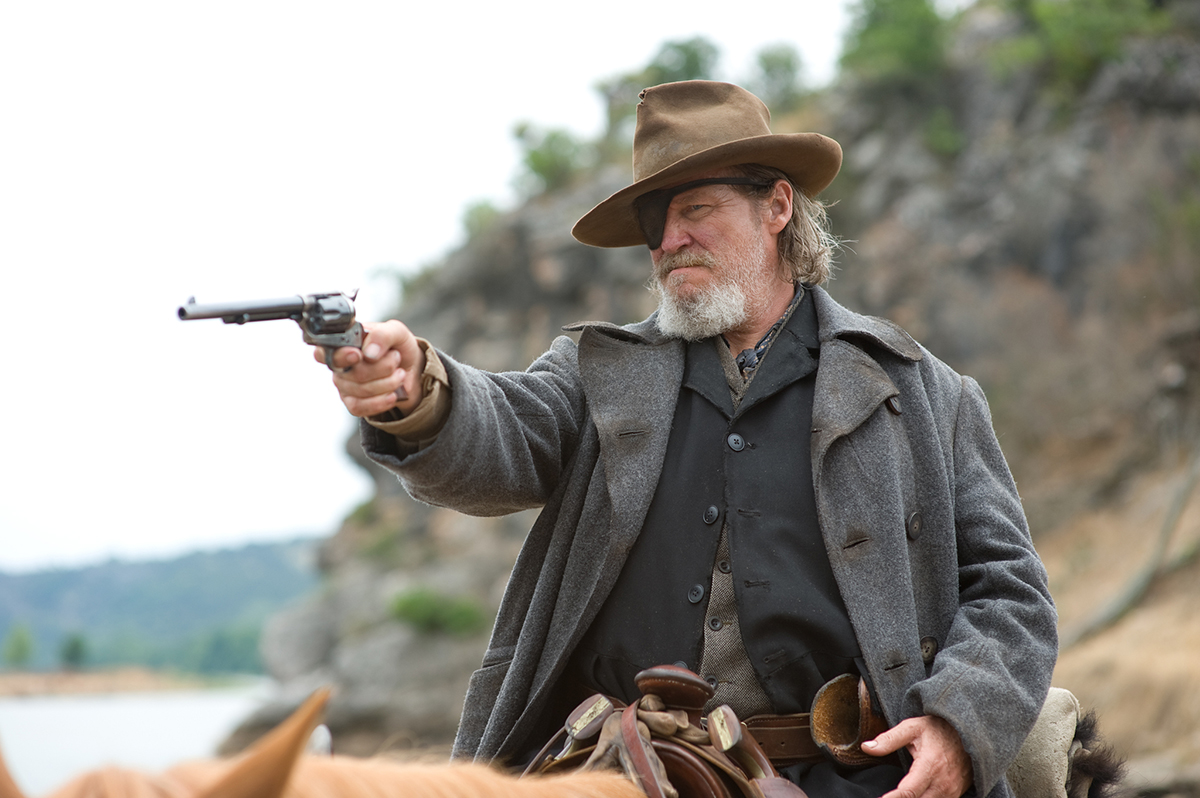 Jeff Bridges with a gun on the back of a horse dressed like a cowboy