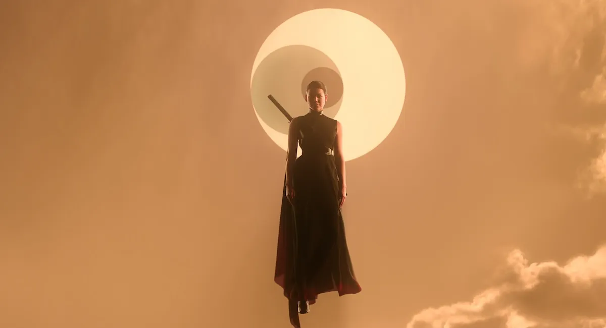 A woman in a black dress with a sword on her back floats in an orange sky, with a solar eclipse behind her.