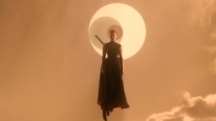 A woman in a black dress with a sword on her back floats in an orange sky, with a solar eclipse behind her.