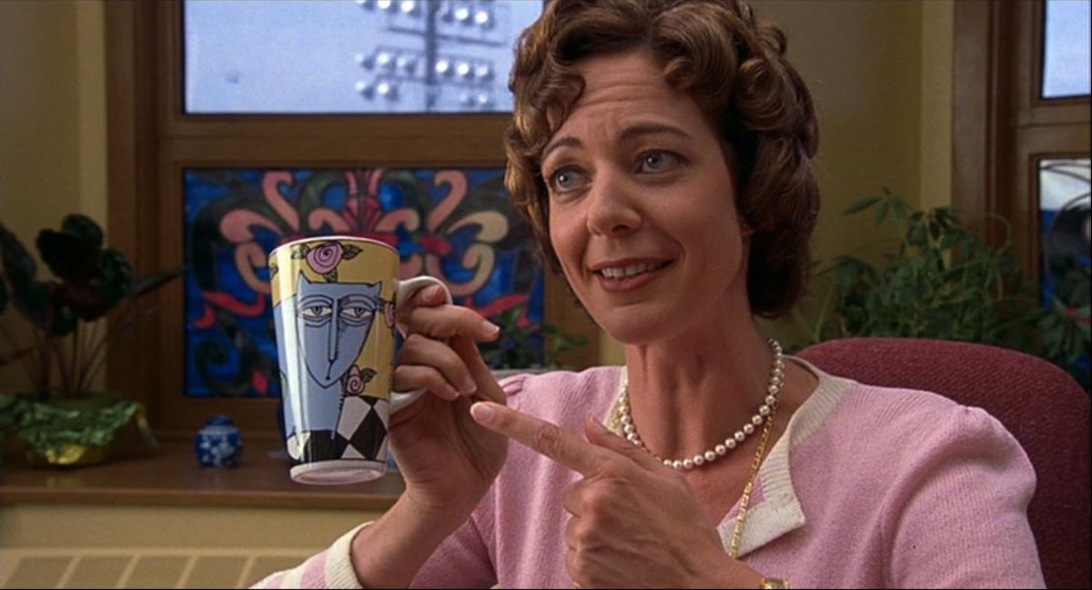 Image of Allison Janney as Ms. Perky in '10 Things I Hate About You.' She is a white woman with curly brown hair sitting at a desk in her office wearing pearls, a pink knit suit, and holding a mug with a cat on it.