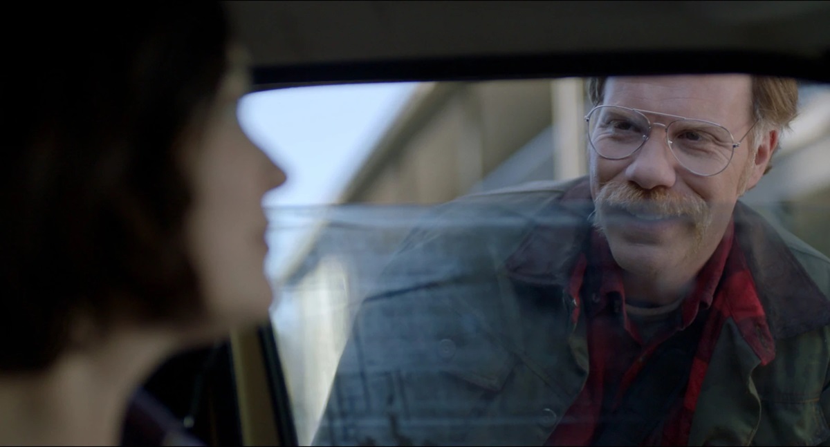 A man with a mustache and glasses peers into a car window at a woman inside