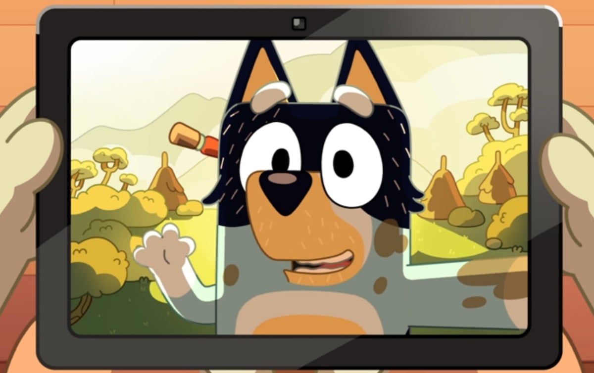 Bandit waves from the screen of a tablet, covered in dirt with a natural landscape behind him.