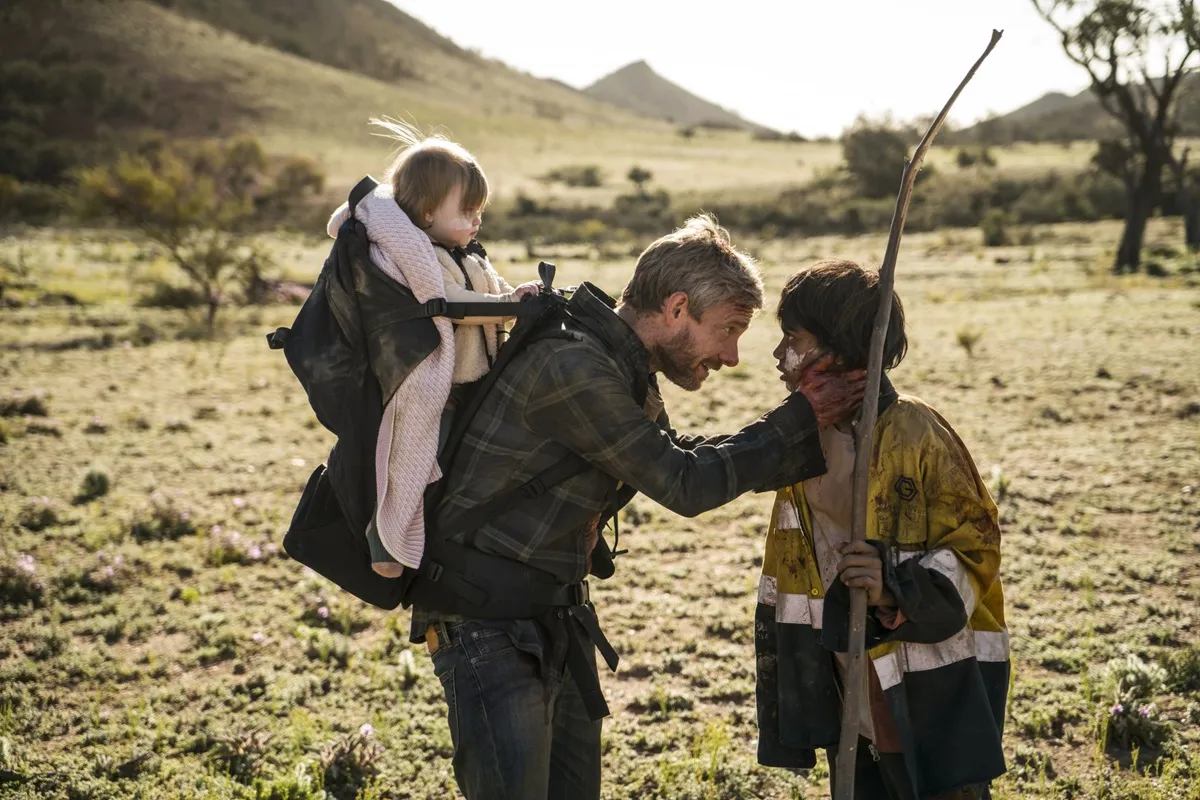 a man (Martin Freeman) wearing an infant carrier backpack crouches to speak to a young boy. 