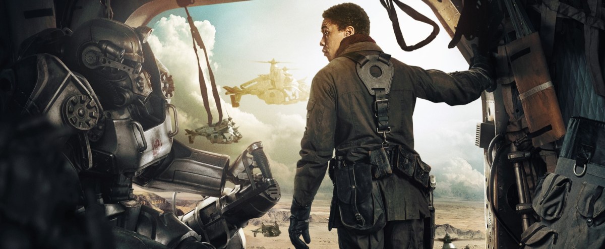 Cropped promotional image of Aaron Moten as Maximus in 'Fallout.' Maximus is a young Black man with short hair wearing a Brotherhood of Steel uniform and standing in the open side of a flying vertibird. He is looking over his shoulder at someone in power armor who's sitting in the cabin with him.