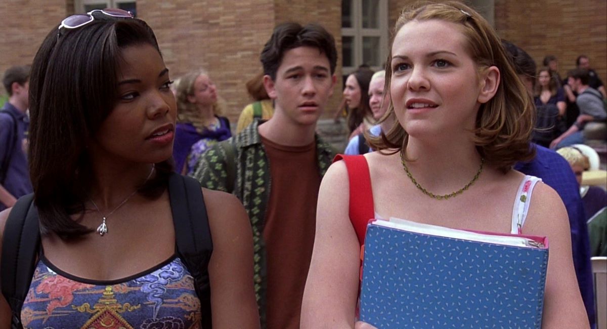 Image of Gabrielle Union as Chastity, Joseph Gordon-Levitt as Cameron, and Larisa Oleynik as Bianca in '10 Things I Hate About You.' Chastity is a Black teenage girl with shoulder length straight hair wearing a patterned tank top and sunglasses on her head. Bianca is a white teenage girl with shoulder length red hair wearing a white sundress and holding a binder to her chest as they walk. Walking behind them, Cameron is a white teenage boy with short dark hair wearing an open green buttondown over a maroon t-shirt.