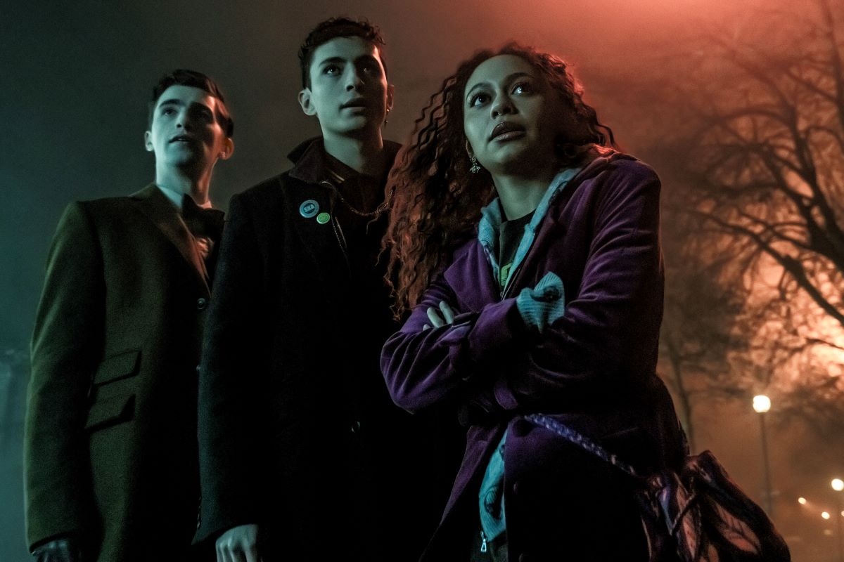 Image of George Rexstrew as Edwin, Jayden Revri as Charles, and Kassius Nelson as Crystal in Netflix's 'Dead Boy Detectives.' Edwin is a white teenager wearing an 1800s boys' school uniform with a bow tie. Charles is a mixed race Indian teenager wearing a 1980s style leather jacket. Crystal is a Black teenager with long, wavy dark hair wearing a purple velvet blazer. They are standing together at night looking off into the distance.