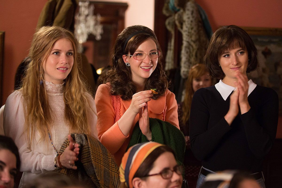 Three young women dressed in the 1960's stand next to each other in a crowd, clapping.