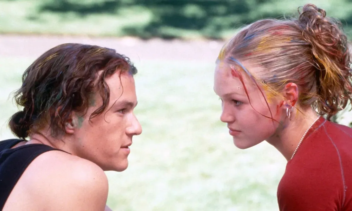 Image of Heath Ledger as Patrick and Julia Stiles as Kat in a scene from '10 Things I Hate About You.' They are both white teenagers sitting on steps staring into each others' eyes. Patrick has wavy, chin-length brown hair and is wearing a black tank top. His hair's pulled back into a short pony tail and there is green paint in his hairline. Kat has wavy blonde hair pulled up into a ponytail, and has various colors of paint in her hair. She's wearing a red shirt.
