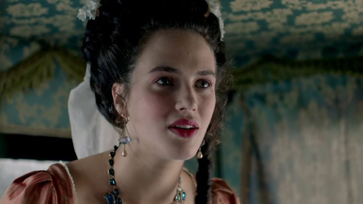 Image of Jessica Brown Findley as Charlotte in Hulu's 'Harlots.' She is a young white woman. Her hair is styled in an up-do that's in fashion for the mid 1700s. She's wearing dangly earrings and wearing pink lipstick and a green jeweled necklace and a pink dress. She's looking ahead in a close-up. 