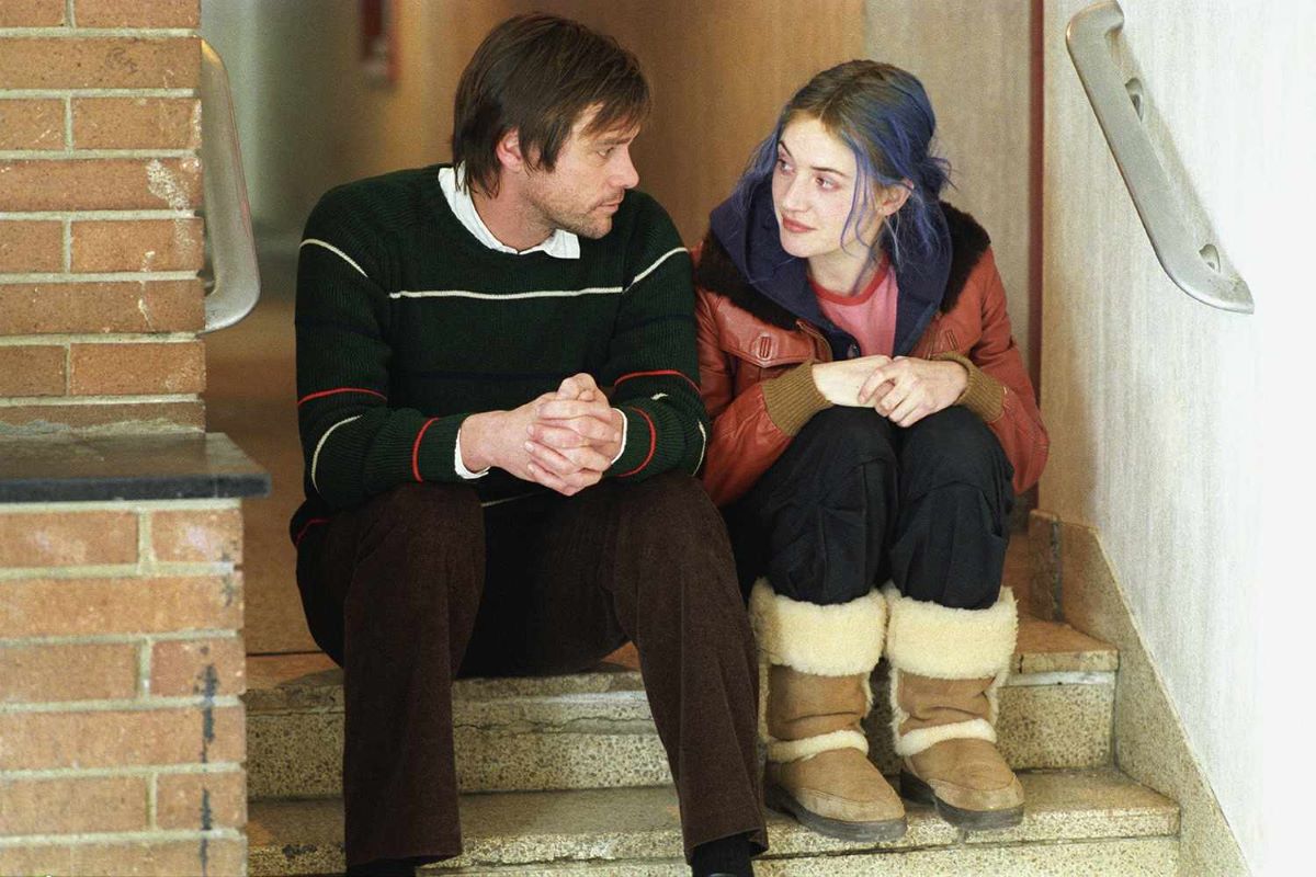 Jim Carrey as Joel and Kate Winslet as Clementine in a scene from "Eternal Sunshine of the Spotless Mind." They are sitting on steps in an apartment building hallway. Joel is a white man with dark hair wearing a green pullover and brown pants. Clementine is a white woman with dyed blue hair wearing a red jacket over a blue hoodie and a red t-shirt with black pants and Ugg boots. 