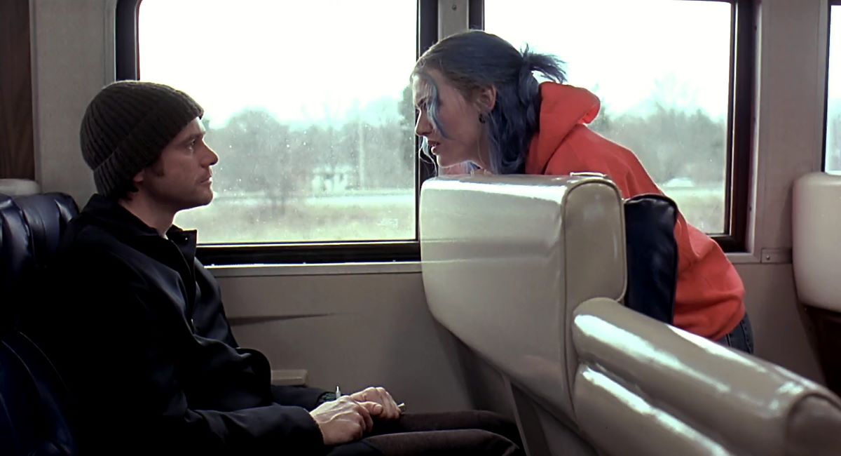 Jim Carrey as Joel and Kate Winslet as Clementine in 'Eternal Sunshine of the Spotless Mind.' They are sitting in separate rows on a Long Island Railroad train, with Clementine turned around on her seat to face him. Joel is a white man with brown hair wearing a black beanie and a black jacket. Clementine is a white woman with dyed blue hair and wearing an orange hoodie. 