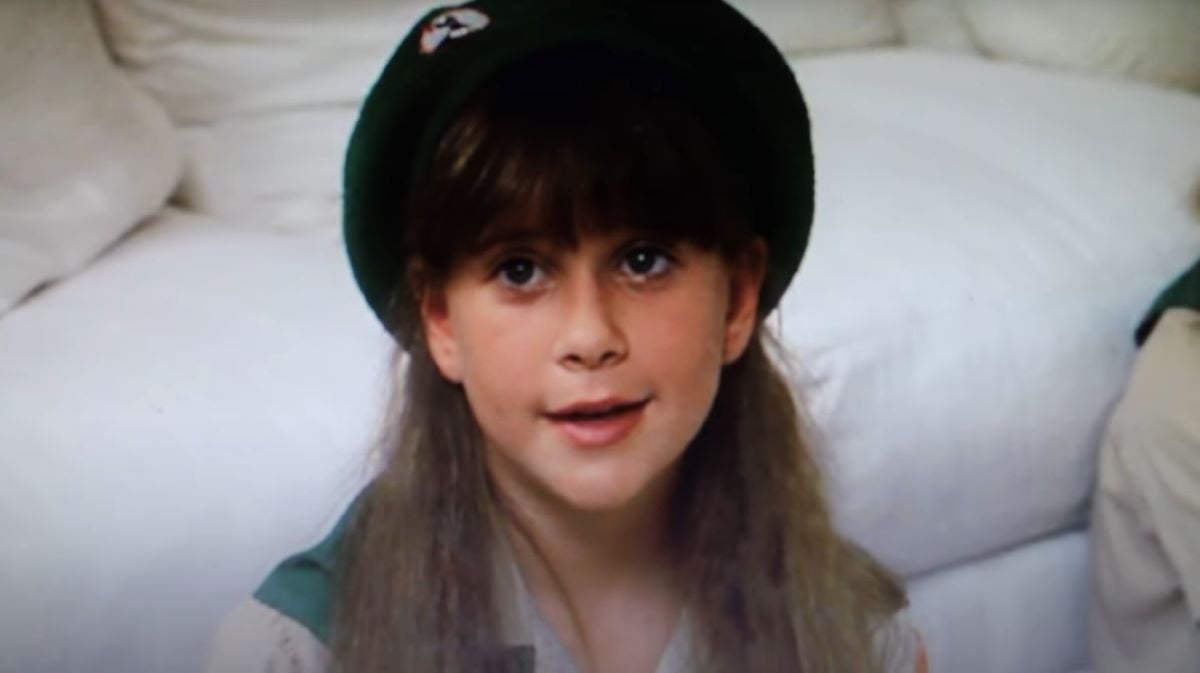 Kellie Martin as Emily in 'Troop Beverly Hills.' She is a white girl with long dark hair and bangs wearing a Wilderness Girls beret. She's sitting on the floor in front of a white couch.