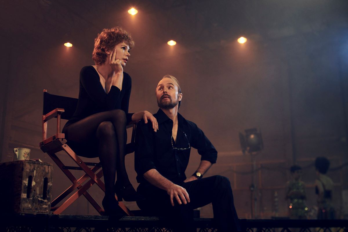 Michelle Williams as Gwen Verdon and Sam Rockwell as Bob Fosse in FX's 'Fosse/Verdon.' Verdon is a white woman with short, curly red hair wearing a long-sleeved black leotard, black stockings, and black dance shoes sitting cross-legged in a directors chair. Fosse is a white man with dark hair and a receding hairline and a beard wearing all black and sitting on the edge of a stage beside Verdon's chair. 