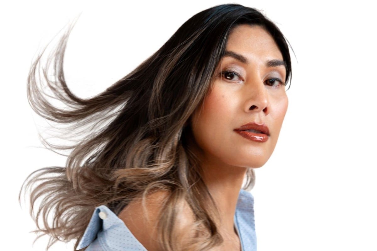 Headshot of Rain Valdez. She is a brown Asian trans woman with long dark hair with blonde highlights blowing in the breeze. Her body is turned to the side with her face turned to the camera. She's wearing a light blue buttondown that's open at the top and coming off her shoulder.