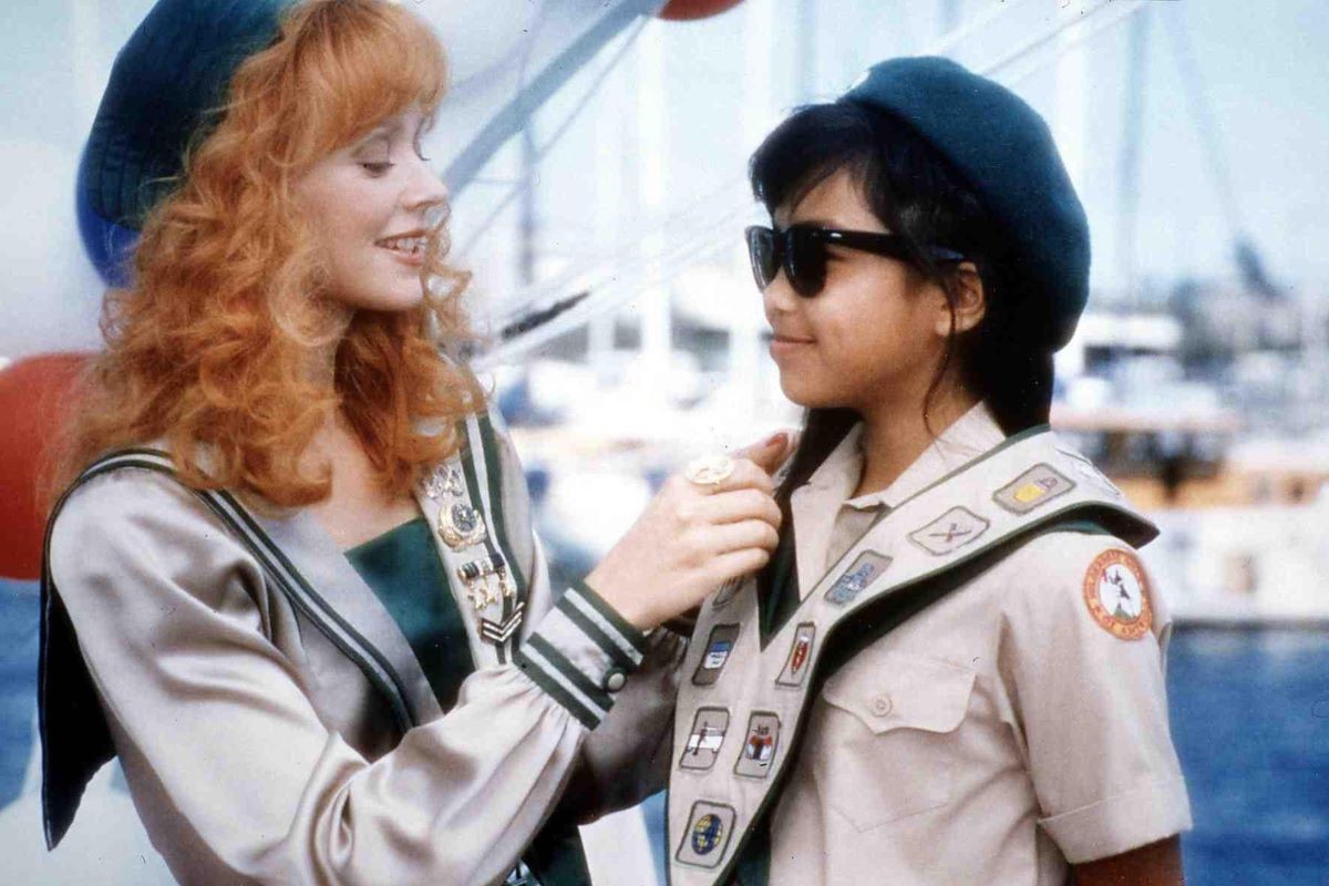 Shelley Long as Phyllis and Aquilina Soriano as Lily in a scene from 'Troop Beverly Hills.' Phyllis is a white woman with curly red hair wearing a customized Wilderness Girl uniform. Lily is a Filipina teen girl with long dark hair wearing her customized uniform and sunglasses. Phyllis is pinning a new patch on Lily's sash. 