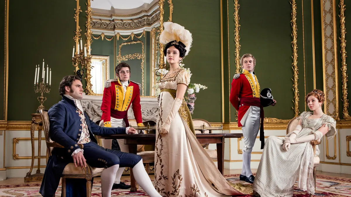 In an opulent Victorian-era room there are two women in cream colored dresses and three men standing around them.