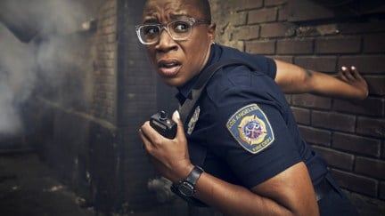Aisha Hinds as Hen with her hand on her walkie and standing by a brick wall