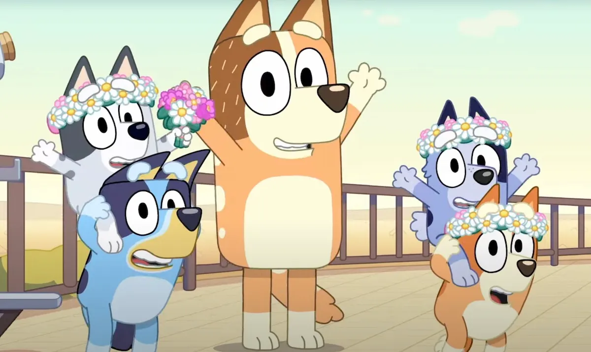 Bluey, Muffin, Socks, Bingo, and Chilli all smile and hold their arms up. The kids are wearing flower crowns.