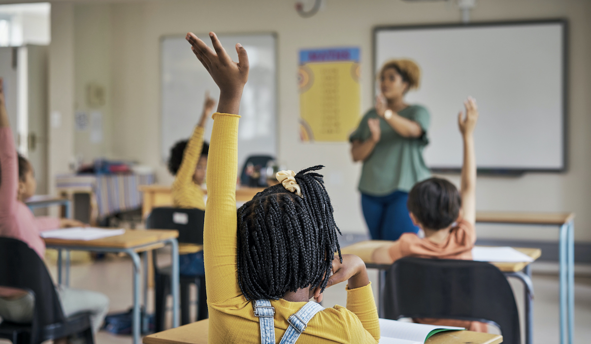 A classroom of young students sitting at their desks, raising hands.
