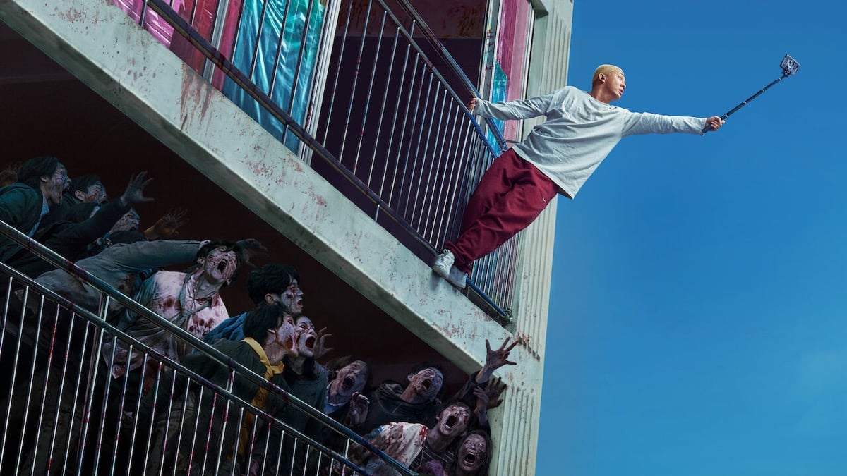A boy hangs out the window of an apartment complex filming zombies with a selfie stick in "#Alive"