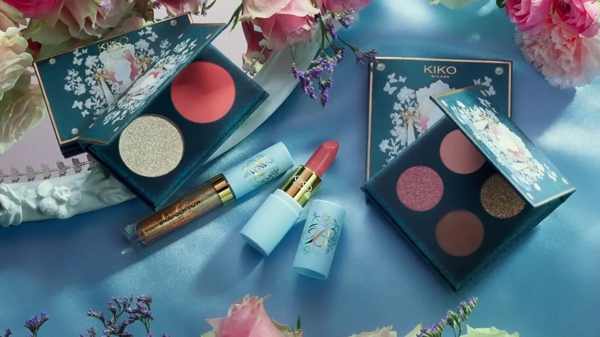 Screencap of the Kiko Milano Bridgerton  make-up collection. There's an open eye palate and blush palate with pinkish/reddish/glittery colors, and an open pinkish lipstick. It's on a blue tablecloth surrounded by pink flowers. 