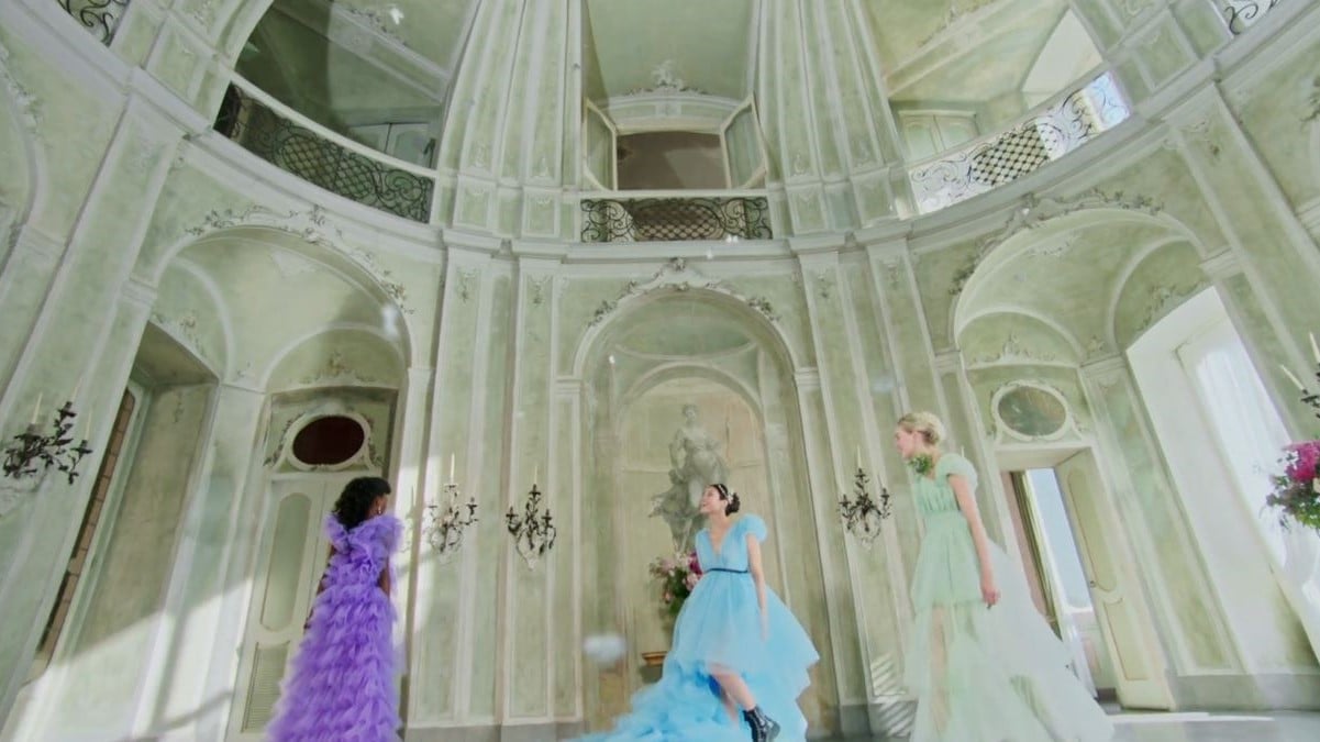 Screencap of three young women in modern, Regency-inspired dresses standing in an ornate hall. The young Black woman is wearing a purple gown. The young Asian woman is wearing a light blue gown and combat boots. And the young white woman is wearing a light green gown. 