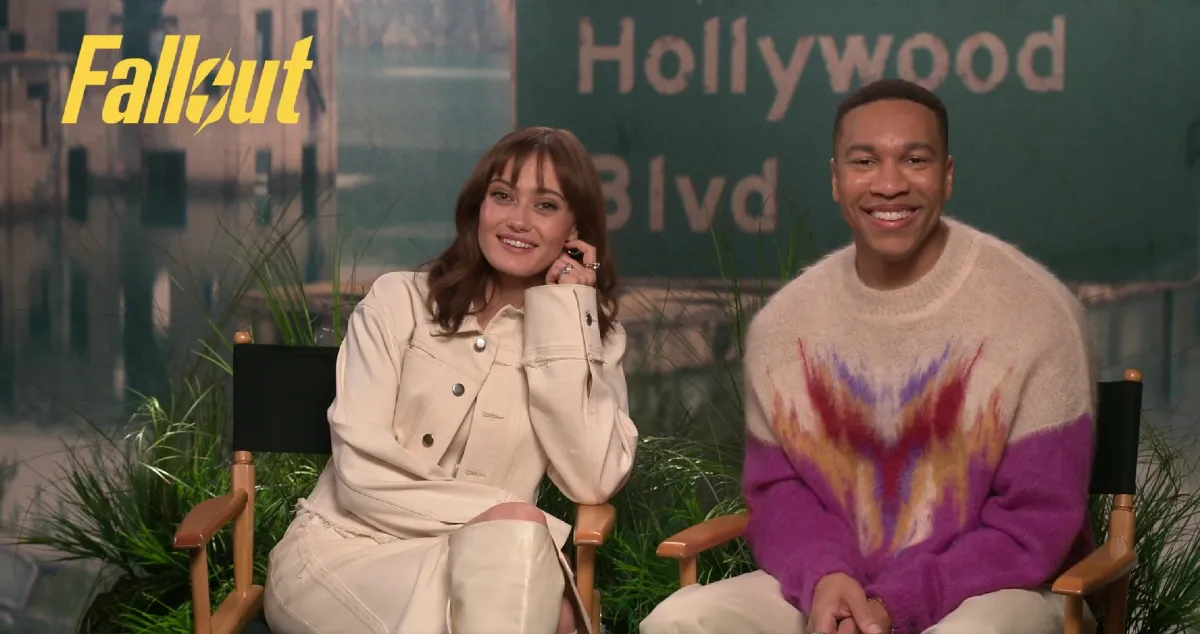 Image of Ella Purnell and Aaron Moten sitting in director chairs during a virtual press junket. Purnell is a white woman with shoulder-length brown hair and wearing an off-white jacket over an off-white dress and knee-high boots. Moten is a Black man with short black hair wearing a sweater that's white at the shoulders with a purple gradient pattern down the middle of it.