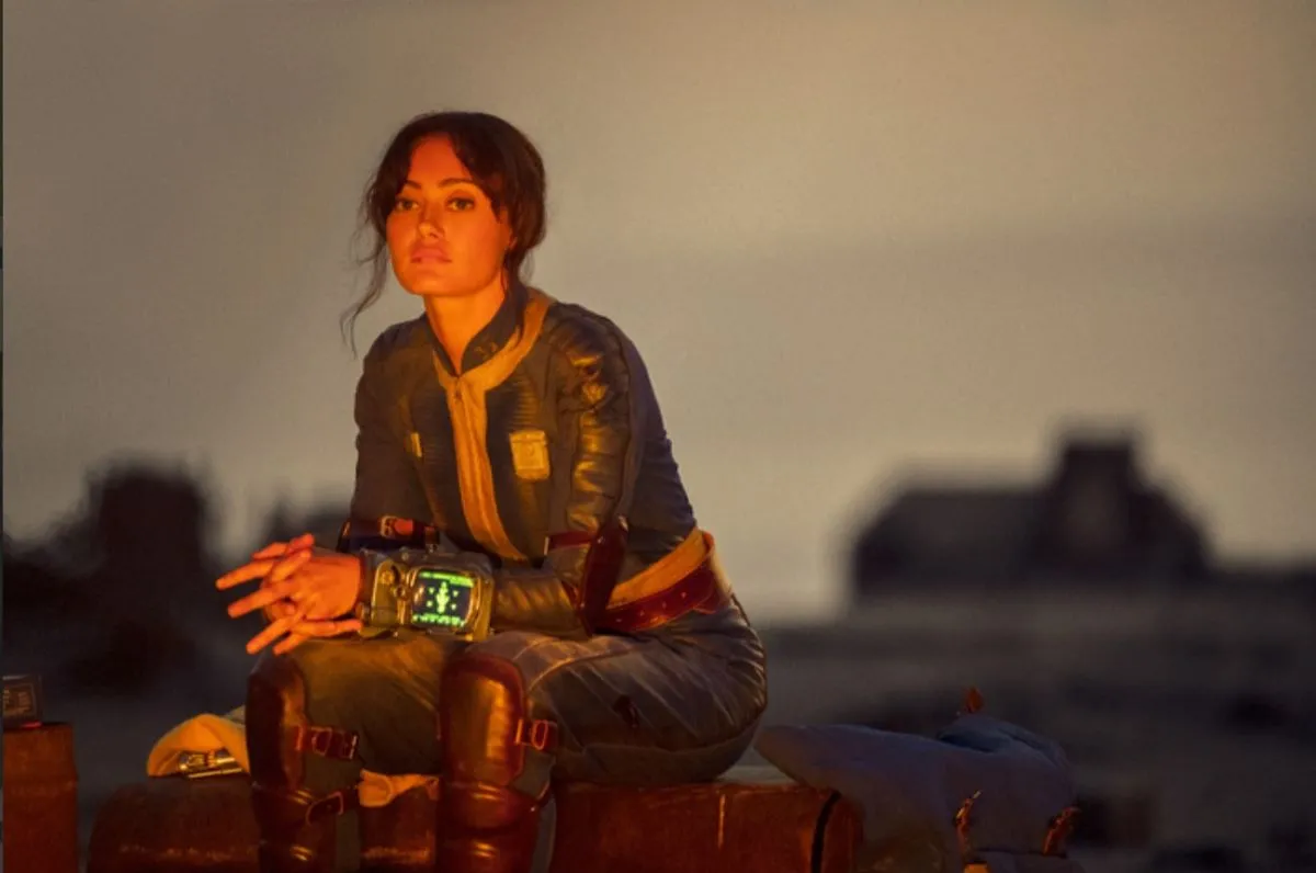 Image of Ella Purnell as Lucy in Prime Video's 'Fallout.' She is a white woman with long, dark hair pulled into a ponytail and wearing a blue 'Fallout' vault suit. She has a Pip-Boy on her wrist and she's sitting on a box in front of a fire at night, resting her arms on her lap as she looks out into the distance.