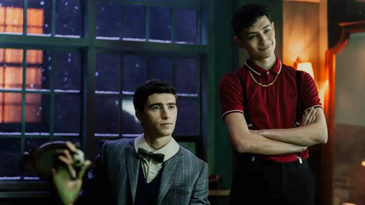 Image of George Rexstrew as Edwin and Jayden Revri as Charles in a scene from Netflix's 'Dead Boy Detectives.' Edwin is a white, teenage boy with short, dark hair wearing a grey pin-striped suit, a white buttondown with a wide collar, and a bowtie who's seated behind a desk with a serious expression. Charles stands beside him with his arms folded. He's a mixed-race white and Indian teenage boy with short, wavy dark hair wearing a maroon polo shirt, a gold chain, and black pants. He's smirking.