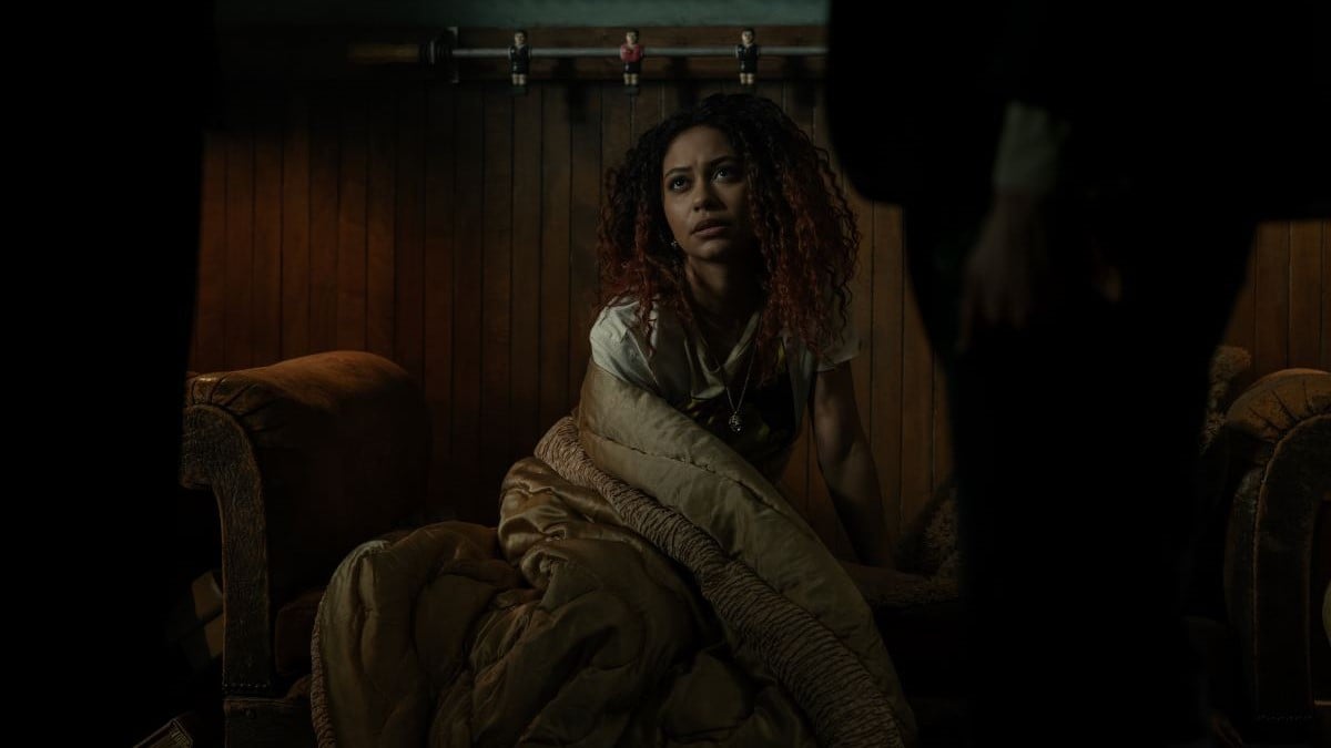Image of Kassius Nelson as Crystal in a scene from Netflix's 'Dead Boy Detectives.' She is a Black teenage girl with shoulder-length, curly black hair highlighted with red wearing a white t-shirt and wrapped in a quilt as she sits up on a couch looking up at the detectives. 