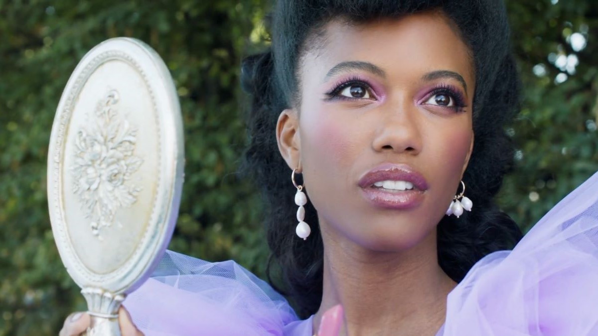 Screencap of a Black female model wearing make-up from the Kiko Milano 'Bridgerton' collection. Her long black hair is pulled back off her face. Her make-up and dress is purple-ish pink, and she's wearing white dangly earrings. She's holding a silver hand mirror and looking off into the distance.