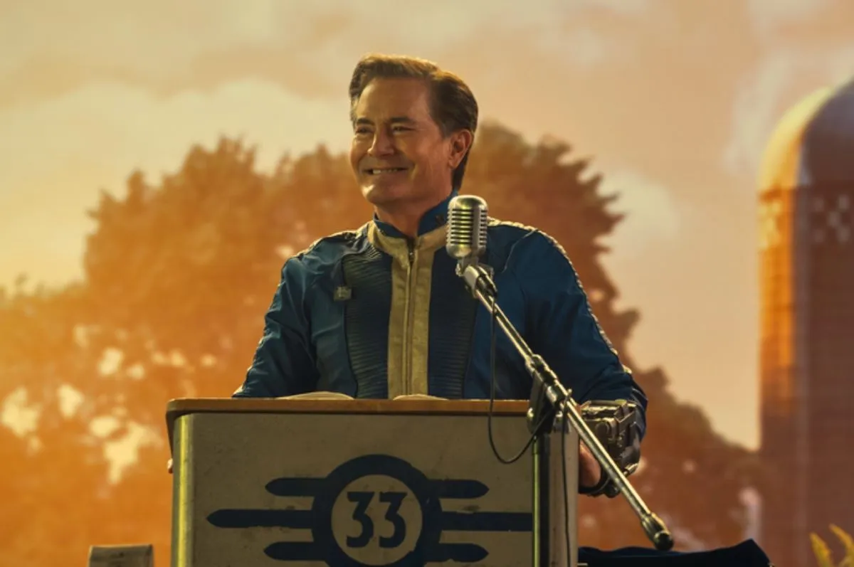 Kyle MacLachlan as Hank MacLean in a scene from Prime Video's 'Fallout.' He is a white man with short dark hair parted on the side wearing a blue and yellow vault suit and standing at a podium with the Vault 33 logo on it. Behind him is a projected image of a sunny farm.
