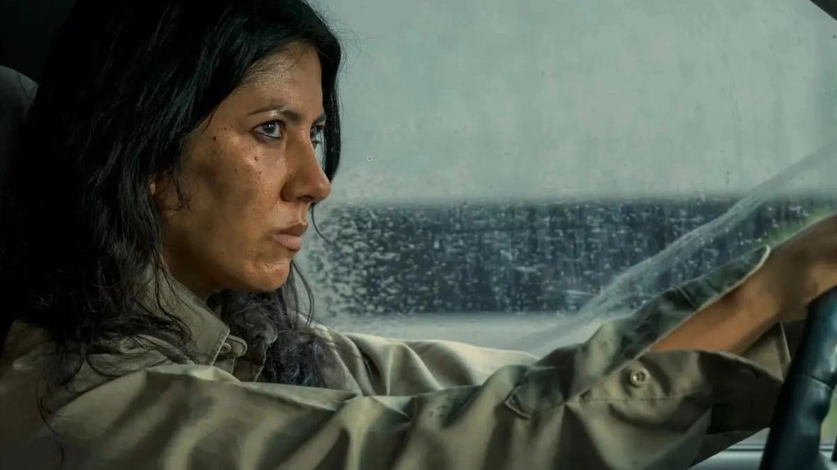 Image of Stephanie Beatriz as Quiet in Peacock's 'Twisted Metal.' She is a brown Latina with long, dark hair wearing an olive green buttondown shirt. She's driving a car in the rain and looking up into the rearview mirror with an angry expression. 