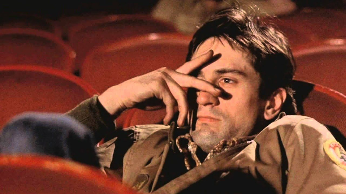 Robert De Niro partially covers his face with his hand while sitting in a movie theater in the film 'Taxi Driver'