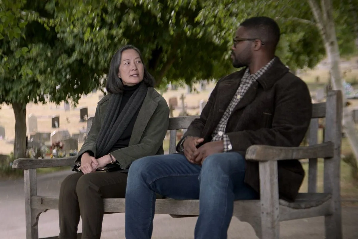 Image of Rosalind Chao as Ye Wenjie and Jovan Adepo as Saul Durand sitting on a park bench in a scene from '3 Body Problem' on Netflix. Ye is a Chinese woman with salt-and-pepper chin length hair wearing a green jacket over a black shirt wearing a dark grey scarf and olive green pants. Saul is a Black man with close-cropped hair and black-rimmed glasses wearing a black coat over a checkered buttondown and jeans.