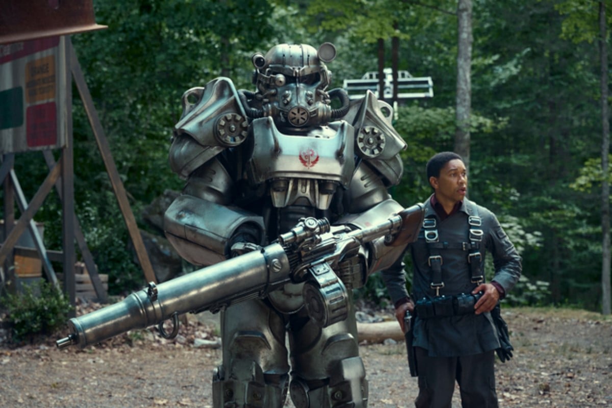 Image of Knight Titus and Maximus (played by Aaron Moten) in a scene from Prime Video's 'Fallout.' Knight Titus is in a suit of silver T-60 power armor with a red Brotherhood of Steel insignia on the chest. He's holding an enormous gun. Maximus is a young Black man with close cropped dark hair wearing a Brotherhood uniform under a parachute harness. They are walking through a wooded area. 