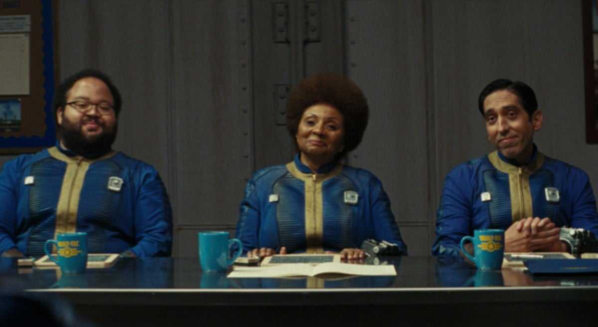 Zach Cherry, Leslie Uggams and Rodrigo Luzzi in a scene from Prime Video's 'Fallout.' All three are seated side-by-side at a long conference table looking out at someone in front of them, and all are wearing blue vault suits. Cherry is a Black man with a short afro and a thick beard and black-rimmed glasses. Uggams is a Black woman with a large afro that's reddish in color. Luzzi