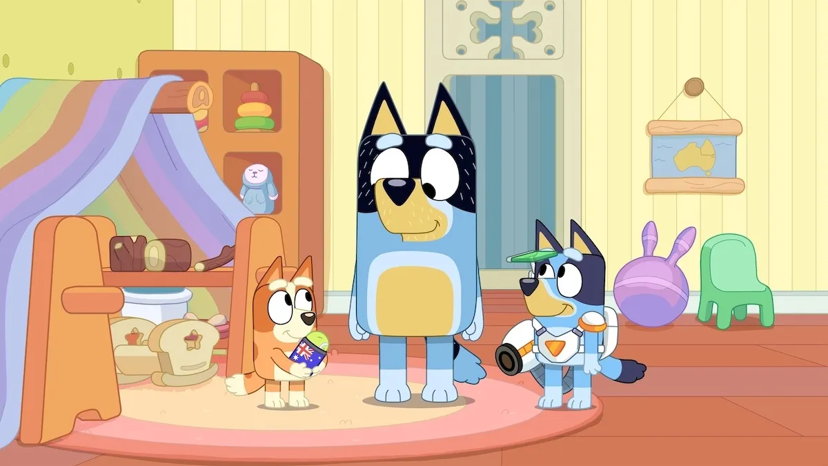 Bandit, Bingo, and Bluey stand together in their playroom. Bluey wears a futuristic cyborg outfit and Bingo holds a tennis ball in a drink sleeve.