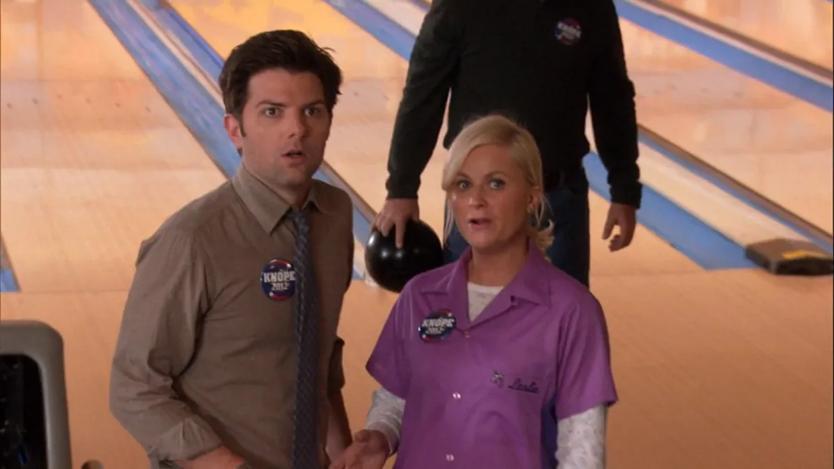 ben and leslie looking shocked while standing in a bowling alley