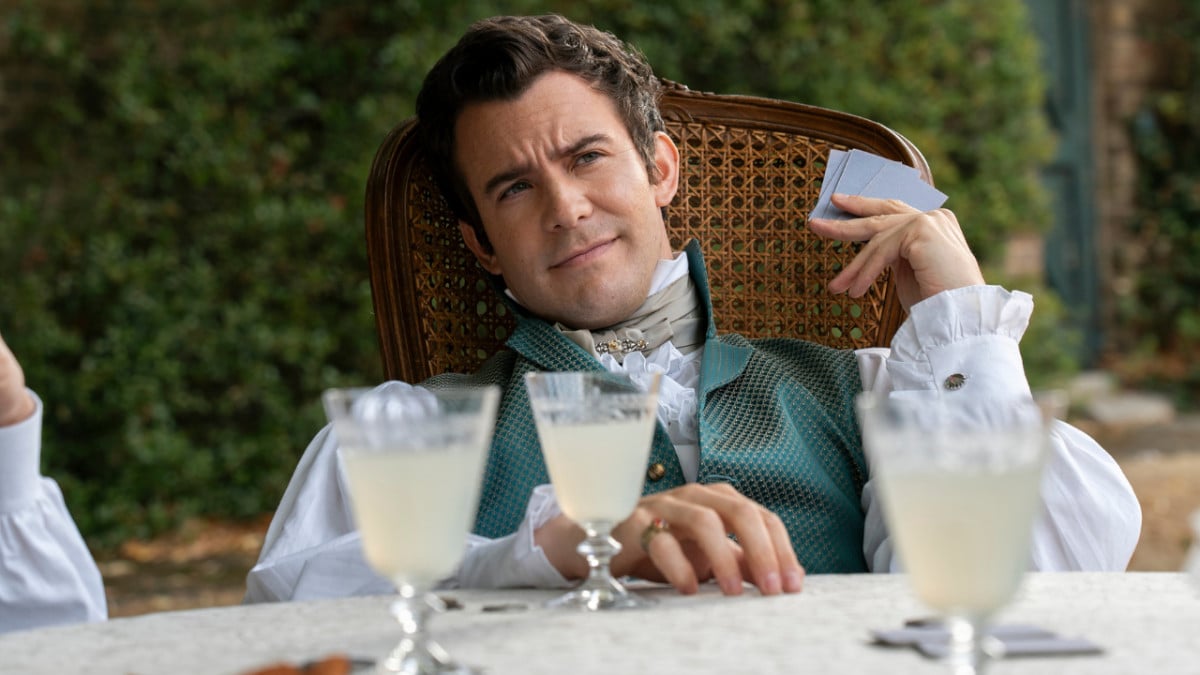 Luke Thompson as Benedict Bridgerton in Bridgerton season 3, slouching in a chair and holing some playing cards. There are drinks on the table in front of him.