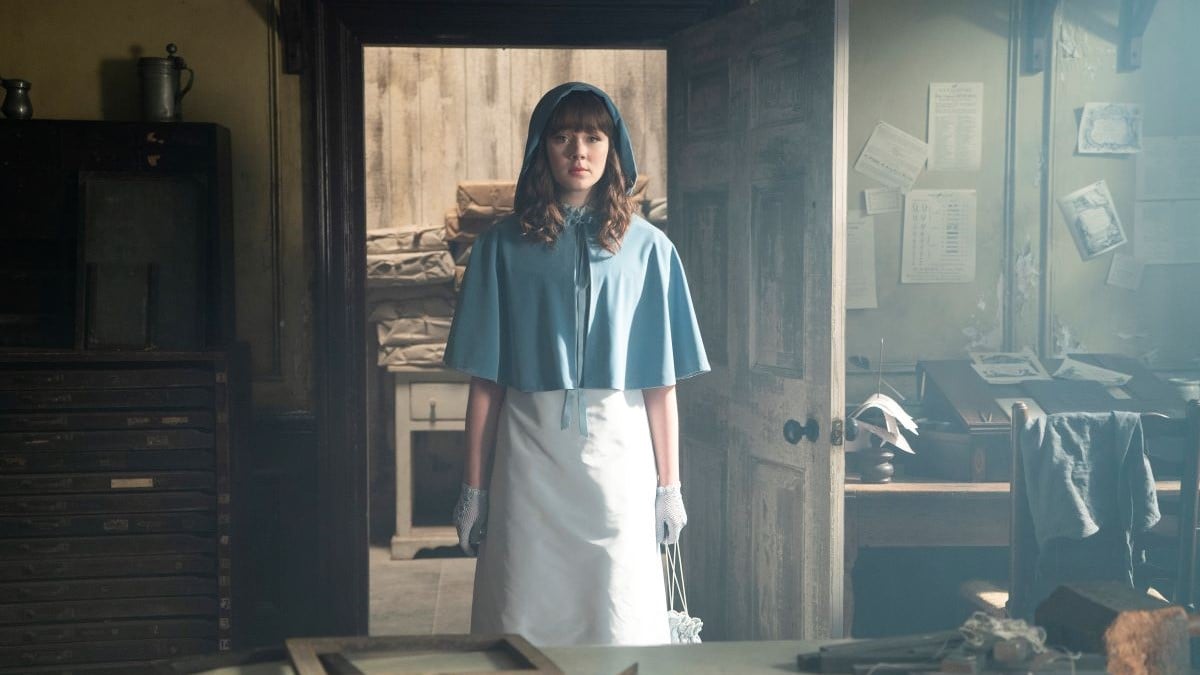 Image of Claudia Jessie as Eloise on Netflix's 'Bridgerton.' She is a young, white woman with long brown hair and bangs. Her hair is down, and she's wearing a light blue hooded cloak and a white dress and carrying a white handbag. She's standing in the doorway of an office.