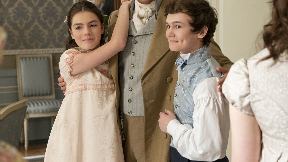 Image of Florence Emilia Hunt as Hyacinth and Will Tilston as Gregory in a scene from Netflix's 'Bridgerton.' They are both white, tween children. Hyacinth has long hair pulled back in a Regency-era style and is wearing a long, light pink dress with cap sleeves. Gregory has short dark hair and is wearing a light blue vest over a white shirt. They are both smiling as they each have an arm around one of their older brothers. 
