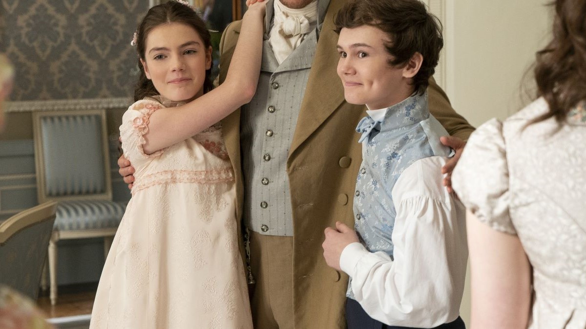 Image of Florence Emilia Hunt as Hyacinth and Will Tilston as Gregory in a scene from Netflix's 'Bridgerton.' They are both white, tween children. Hyacinth has long hair pulled back in a Regency-era style and is wearing a long, light pink dress with cap sleeves. Gregory has short dark hair and is wearing a light blue vest over a white shirt. They are both smiling as they each have an arm around one of their older brothers.
