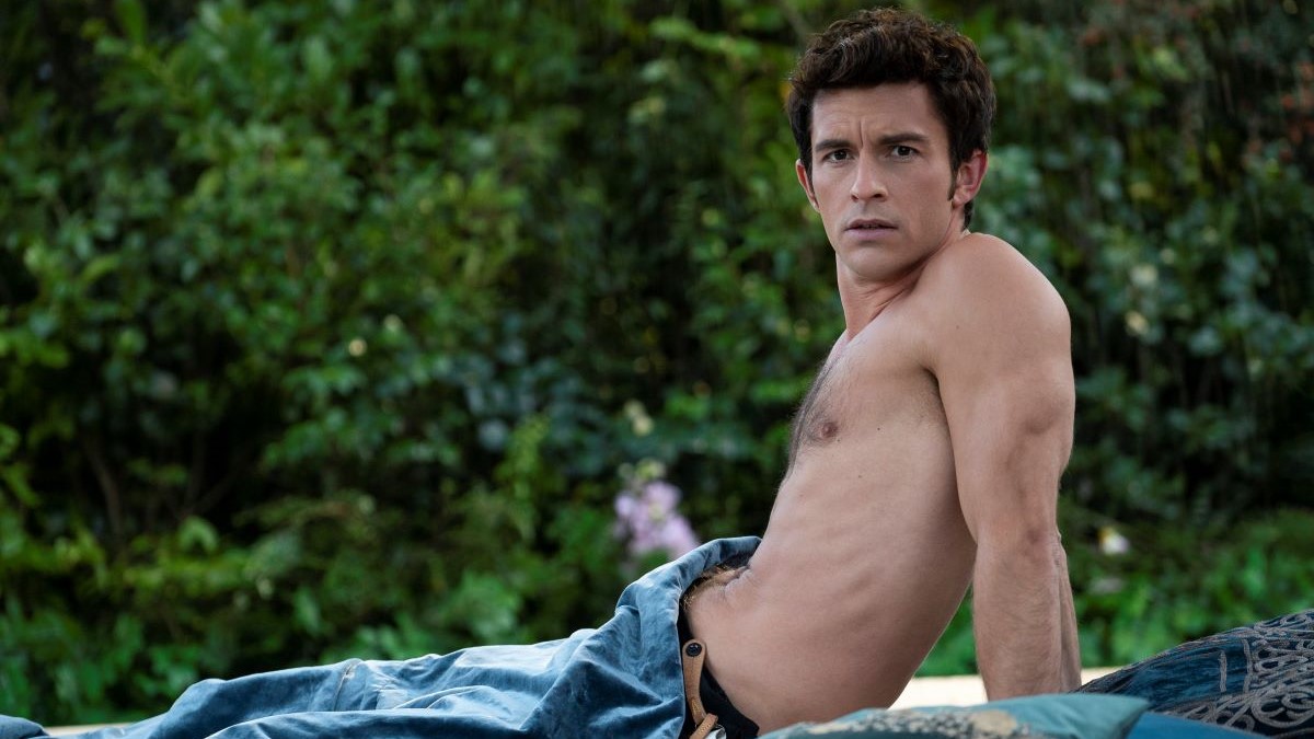 Image of Jonathan Bailey as Anthony in Netflix's 'Bridgerton.' He is a white man with thick dark hair sitting shirtless on a blanket in a garden. He's sitting back, propped up on his arms as he looks at someone off to the side. 